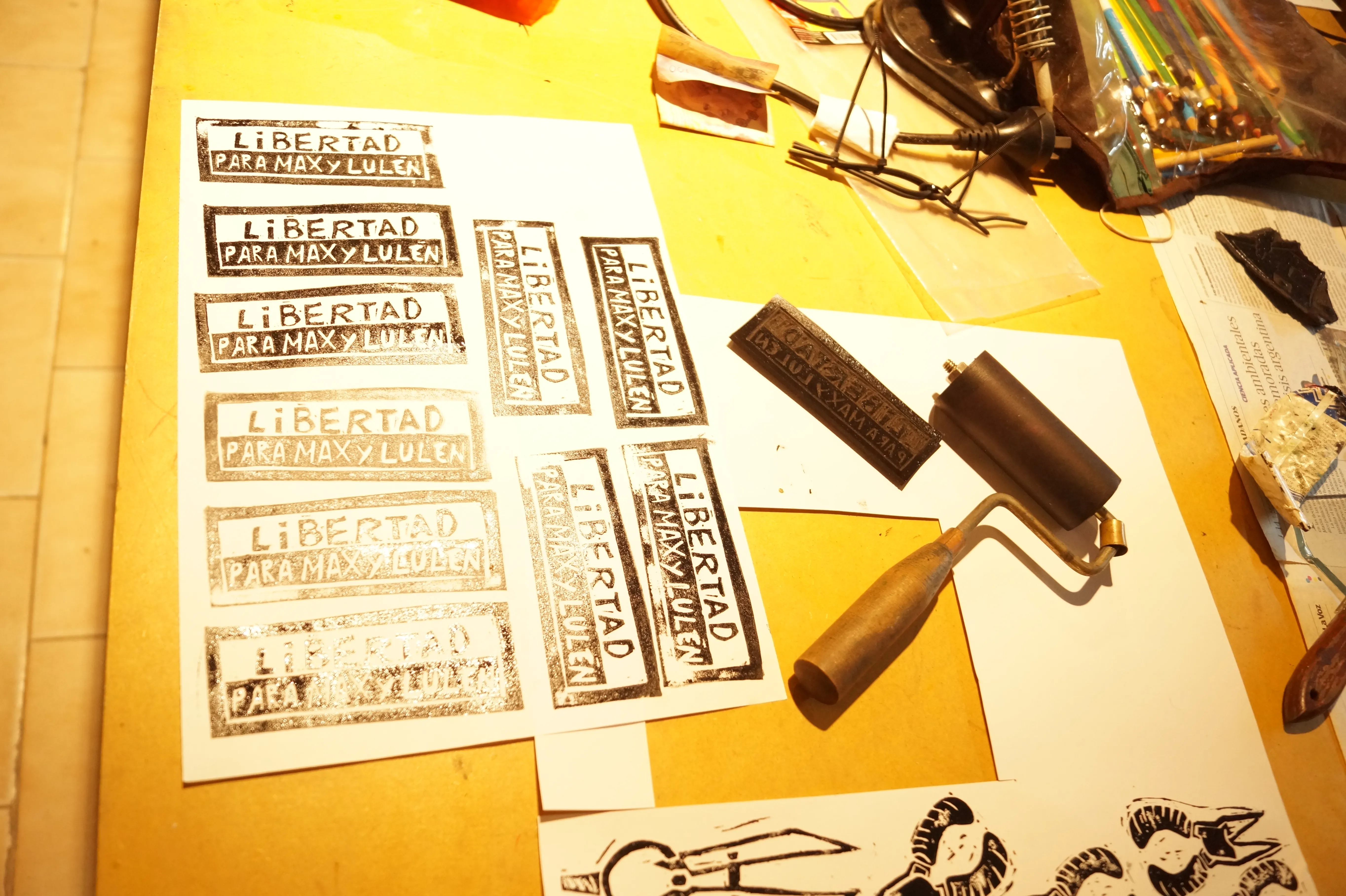 Foto de una mesa de trabajo en donde se ven herramientas de grabado en xilografía y papeles xilografiados con la consigna: Libertad para Max y Lulen